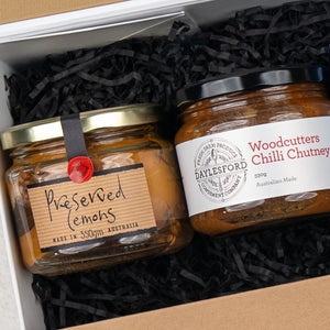 The Four Pillars Botanical Discovery Hamper by The Hamper Boutique Co includes two glass jars: Preserved Lemons with a handwritten tag and Woodcutters Chilli Chutney with a printed label, packed in shredded black paper. Both products are from Australia.