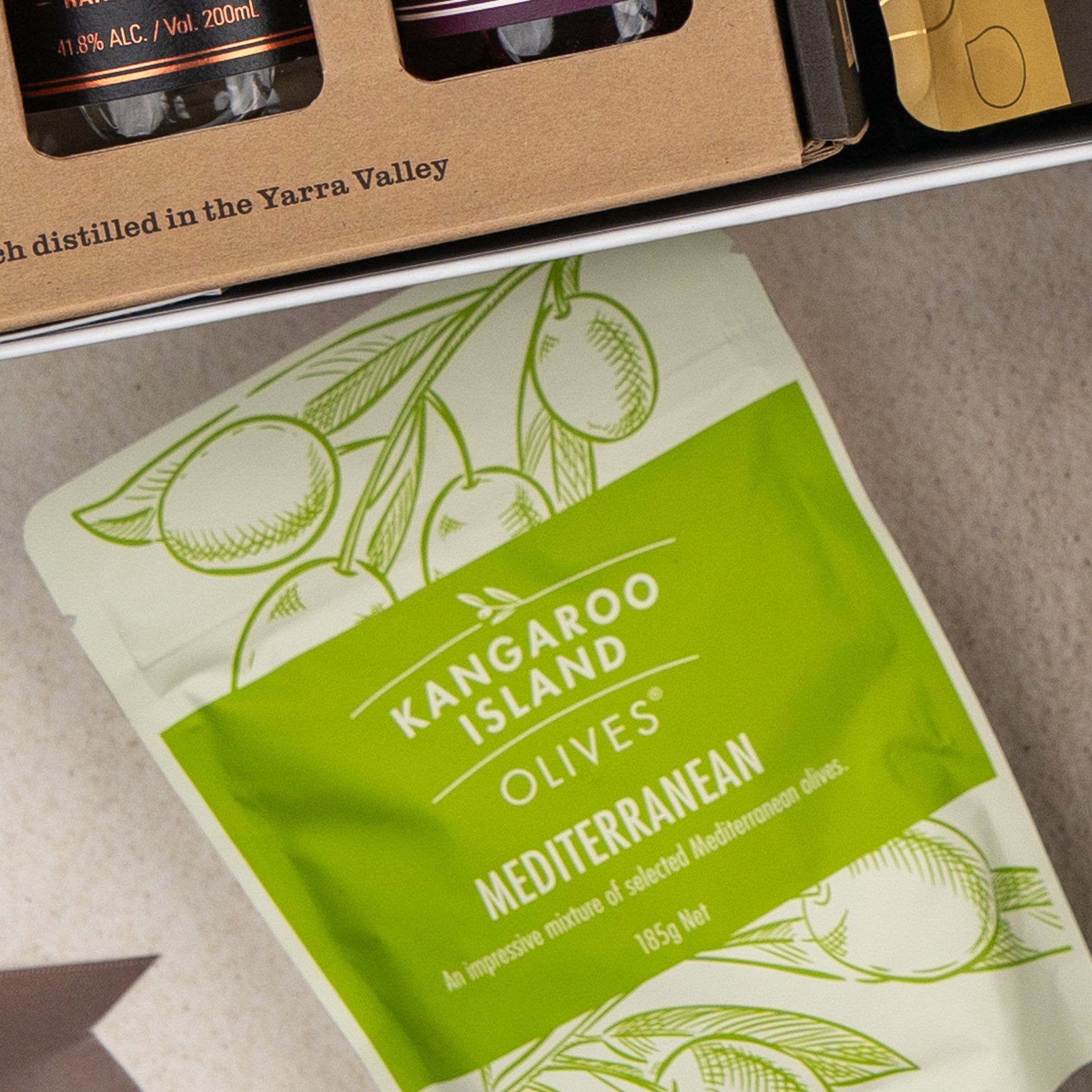 A close-up of a green and white Kangaroo Island Olives Mediterranean pouch sits beside The Hamper Boutique Co's Four Pillars Botanical Discovery Hamper, with glass bottles of amber liquid partially visible at the top.