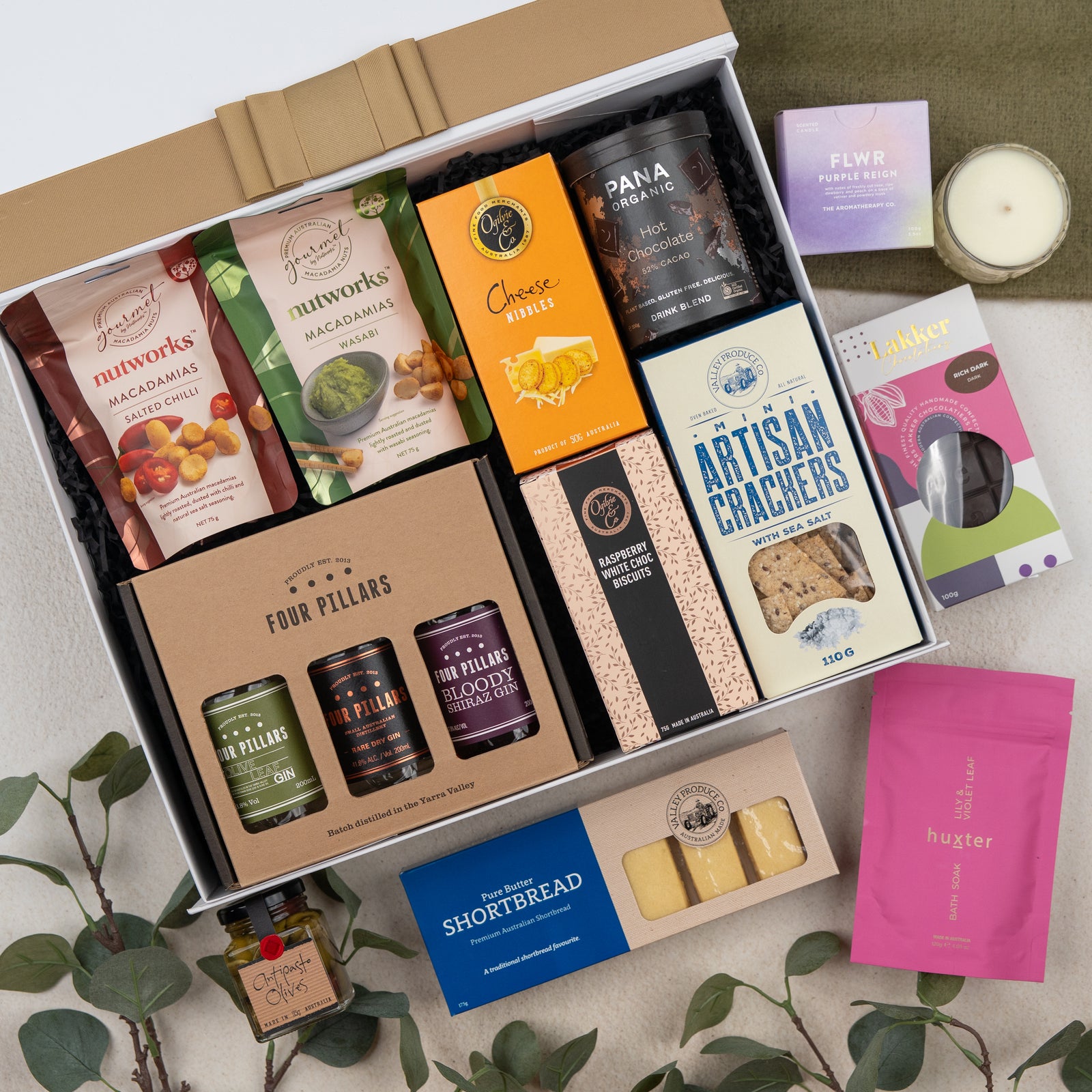 The Hamper Boutique Co’s Four Pillars Ultimate Gin Experience Hamper features gourmet treats like crackers, shortbread, macadamias, cheese nibbles, gin marmalade, plus a candle, tea, and soap—beautifully presented with green leaves.