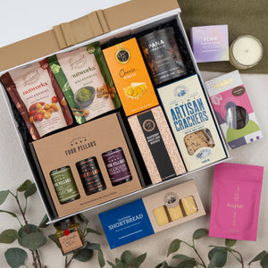 The Hamper Boutique Co’s Four Pillars Ultimate Gin Experience Hamper features gourmet treats like crackers, shortbread, macadamias, cheese nibbles, gin marmalade, plus a candle, tea, and soap—beautifully presented with green leaves.
