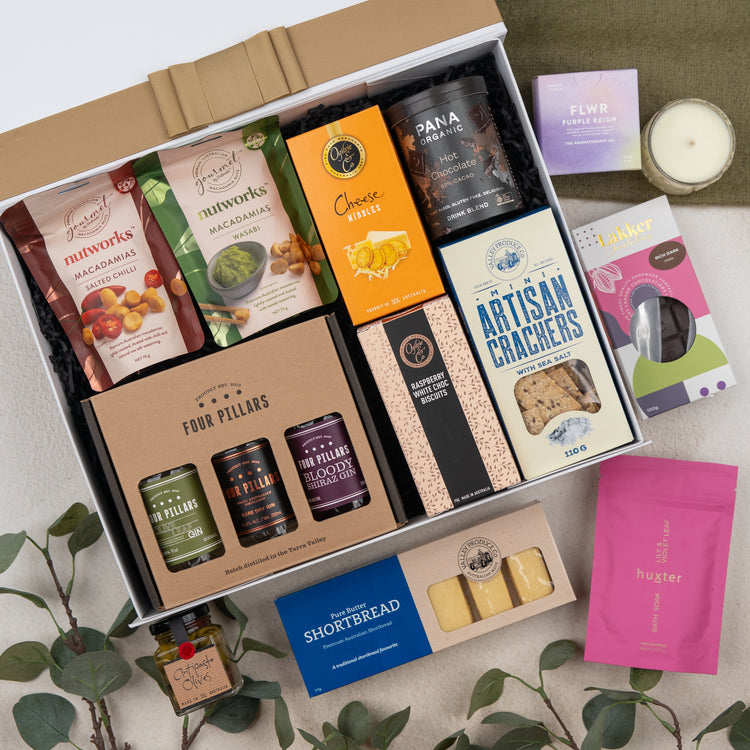 The Hamper Boutique Co’s Four Pillars Ultimate Gin Experience Hamper features gourmet treats like crackers, shortbread, macadamias, cheese nibbles, gin marmalade, plus a candle, tea, and soap—beautifully presented with green leaves.