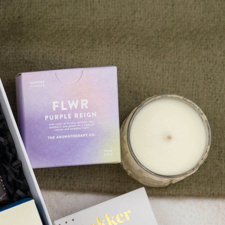 A Four Pillars Ultimate Gin Experience Hamper from The Hamper Boutique Co is displayed on a green surface, accompanied by a white candle in a glass holder. A book and a white box can also be seen in the corner.