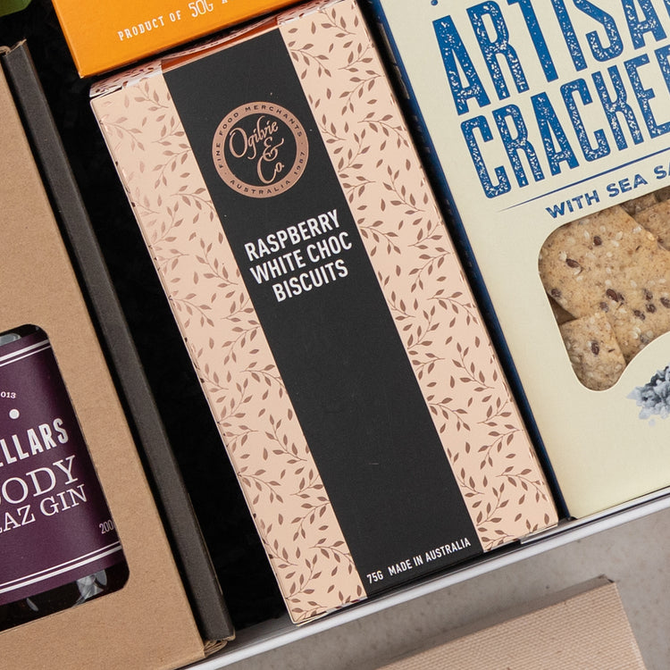 A pink box of Ogilvie & Co Raspberry White Choc Biscuits with a black stripe and leaf pattern is nestled between gin and artisan crackers in The Hamper Boutique Co's Four Pillars Ultimate Gin Experience Hamper.