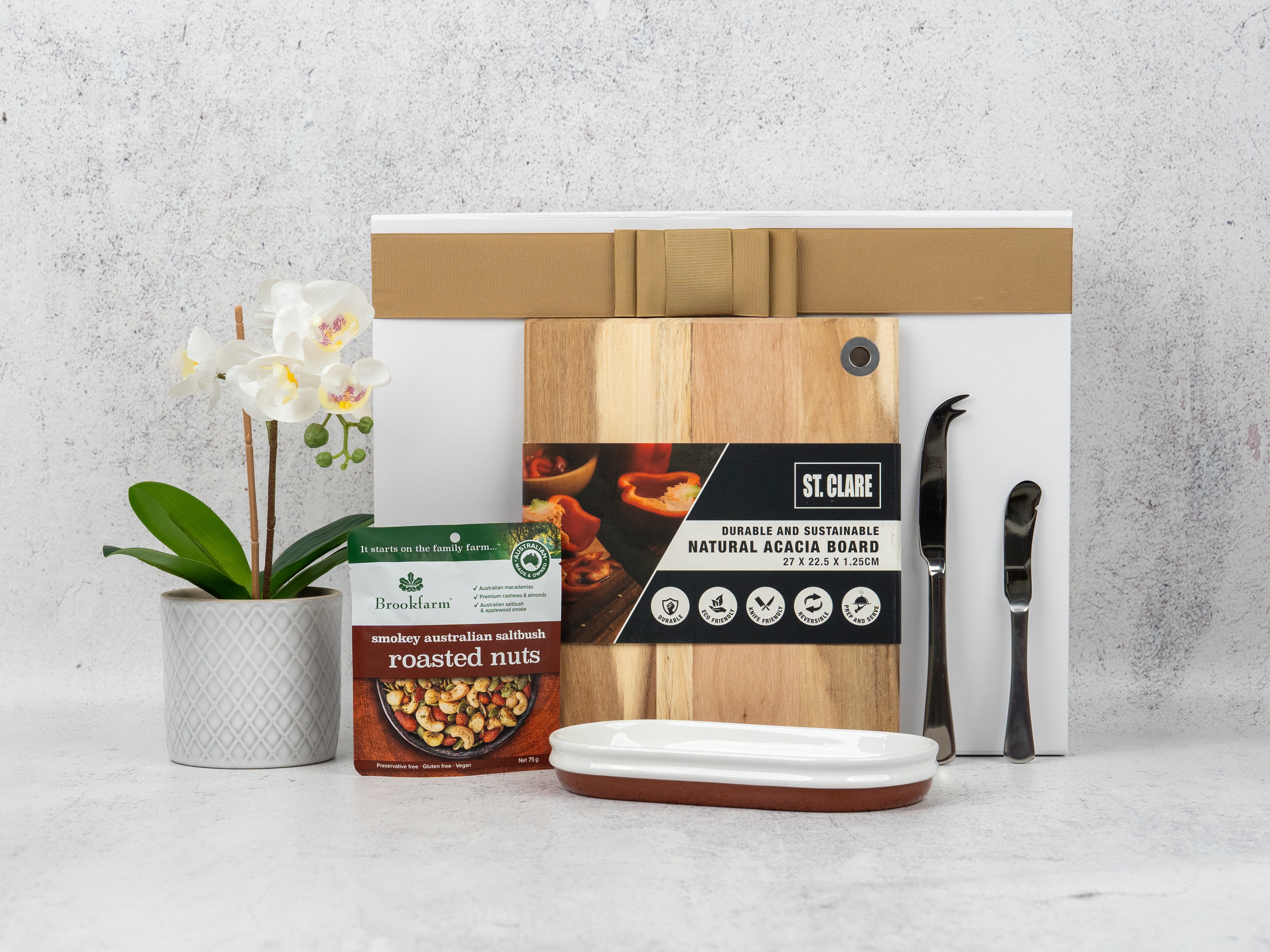 The Hamper Boutique Co’s Artisan Home and Food Hamper features a potted orchid, roasted nuts, an acacia wood cutting board, a white rectangular dish, metal tongs, and two stacked boxes, beautifully displayed on a light surface.