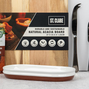 An Artisan Home and Food Hamper by The Hamper Boutique Co features a natural acacia cutting board standing behind a white and brown ceramic dish, with two cheese knives and a partially visible box nearby.
