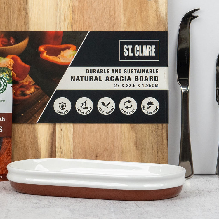 An Artisan Home and Food Hamper by The Hamper Boutique Co features a natural acacia cutting board standing behind a white and brown ceramic dish, with two cheese knives and a partially visible box nearby.