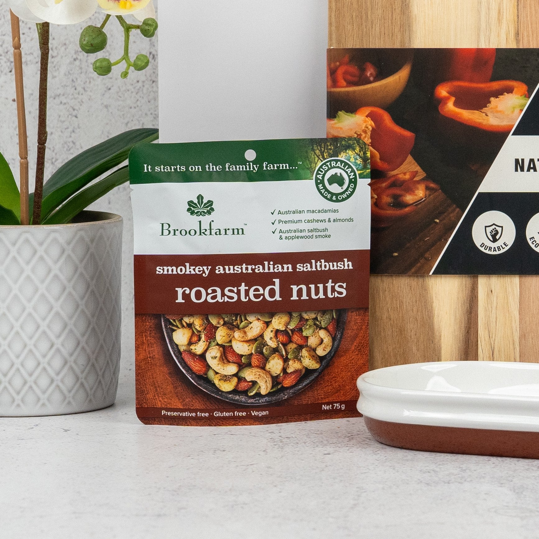 A packet of The Hamper Boutique Co Artisan Home and Food Hamper sits on a kitchen counter beside a wooden cutting board, a white dish, and a potted orchid. The packet features an image of the hamper’s contents on the front.