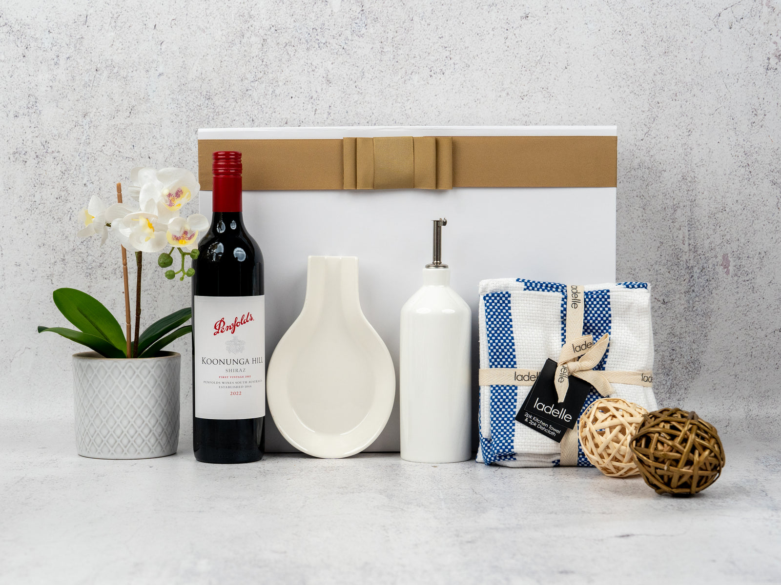 The Penfolds and Homeware gift set by The Hamper Boutique Co features a potted white orchid, red wine, white ceramic dish, ceramic oil dispenser, blue and white kitchen towels, wicker balls, all presented before a ribboned white box.