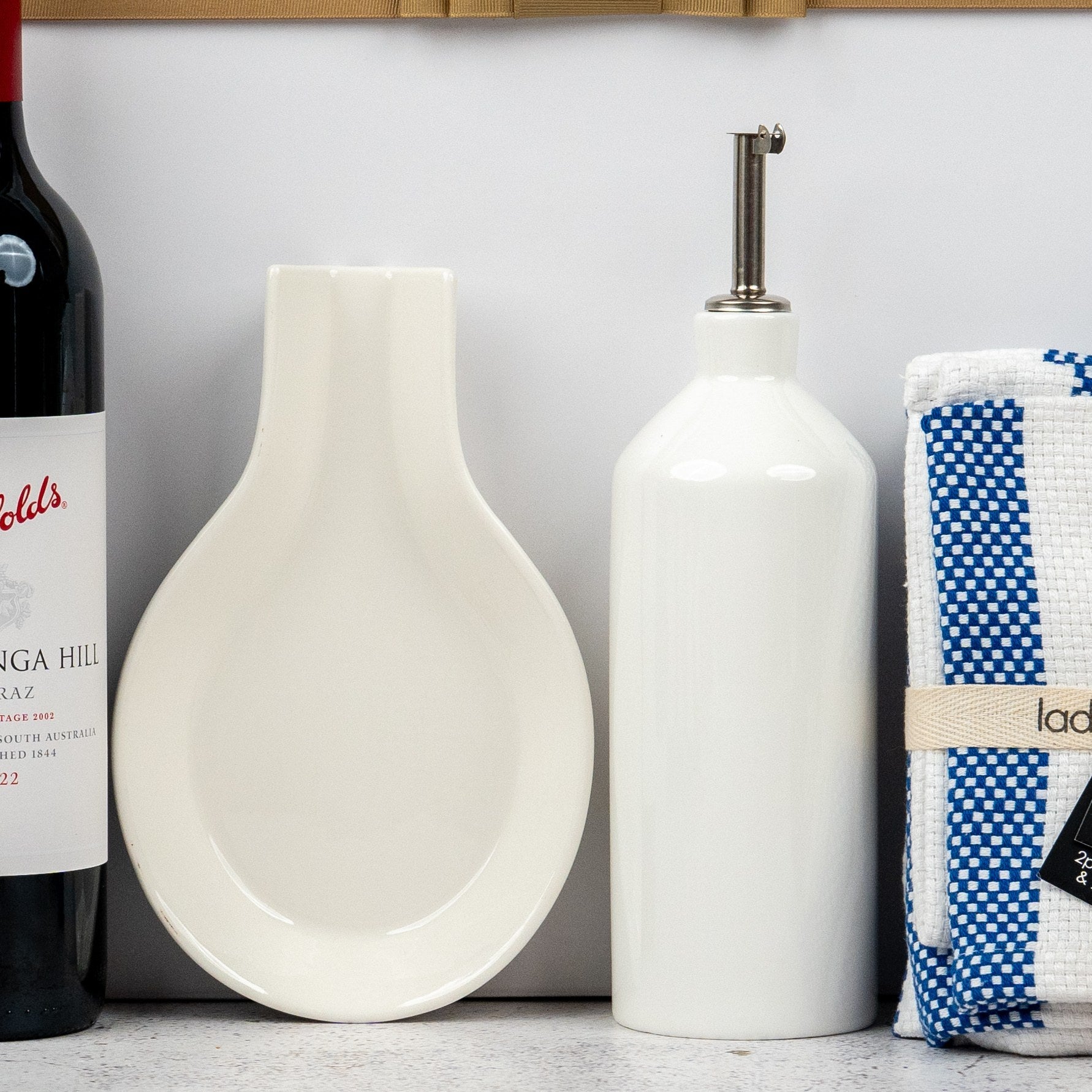 A selection from The Hamper Boutique Co’s Penfolds and Homeware features a red wine bottle, white ceramic spoon rest, white oil dispenser with metal spout, and a folded blue-and-white checkered kitchen towel on the countertop.