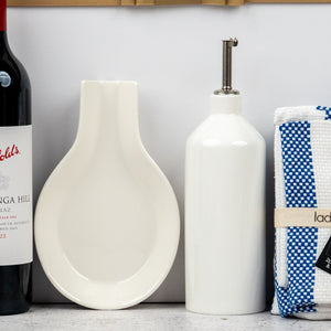 A selection from The Hamper Boutique Co’s Penfolds and Homeware features a red wine bottle, white ceramic spoon rest, white oil dispenser with metal spout, and a folded blue-and-white checkered kitchen towel on the countertop.