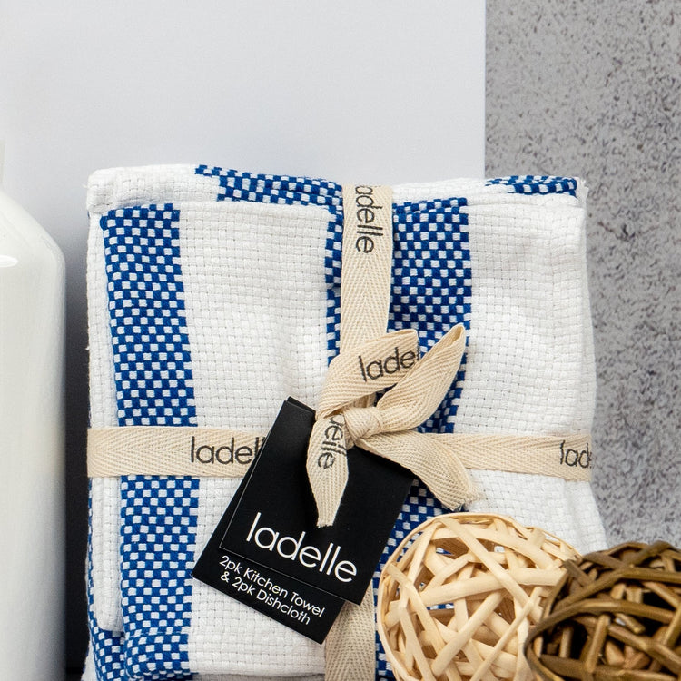 A bundle of blue and white Penfolds and Homeware kitchen towels from The Hamper Boutique Co, tied with a beige ribbon, sits beside decorative wicker balls and a white bottle on a light surface.
