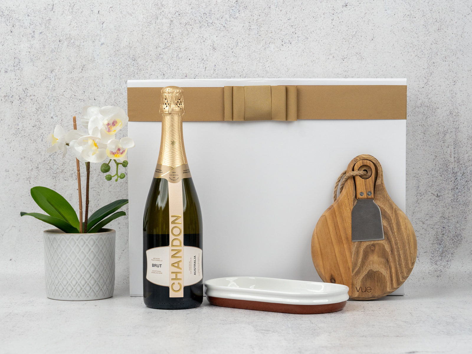 The Hamper Boutique Co’s Sparkling Celebration Hamper includes a bottle of Chandon Brut, white potted orchid, ceramic dish, two wooden cheese boards with a cheese knife in a gold-ribboned white box—all beautifully arranged.