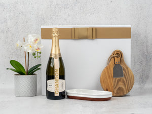 The Hamper Boutique Co’s Sparkling Celebration Hamper includes a bottle of Chandon Brut, white potted orchid, ceramic dish, two wooden cheese boards with a cheese knife in a gold-ribboned white box—all beautifully arranged.