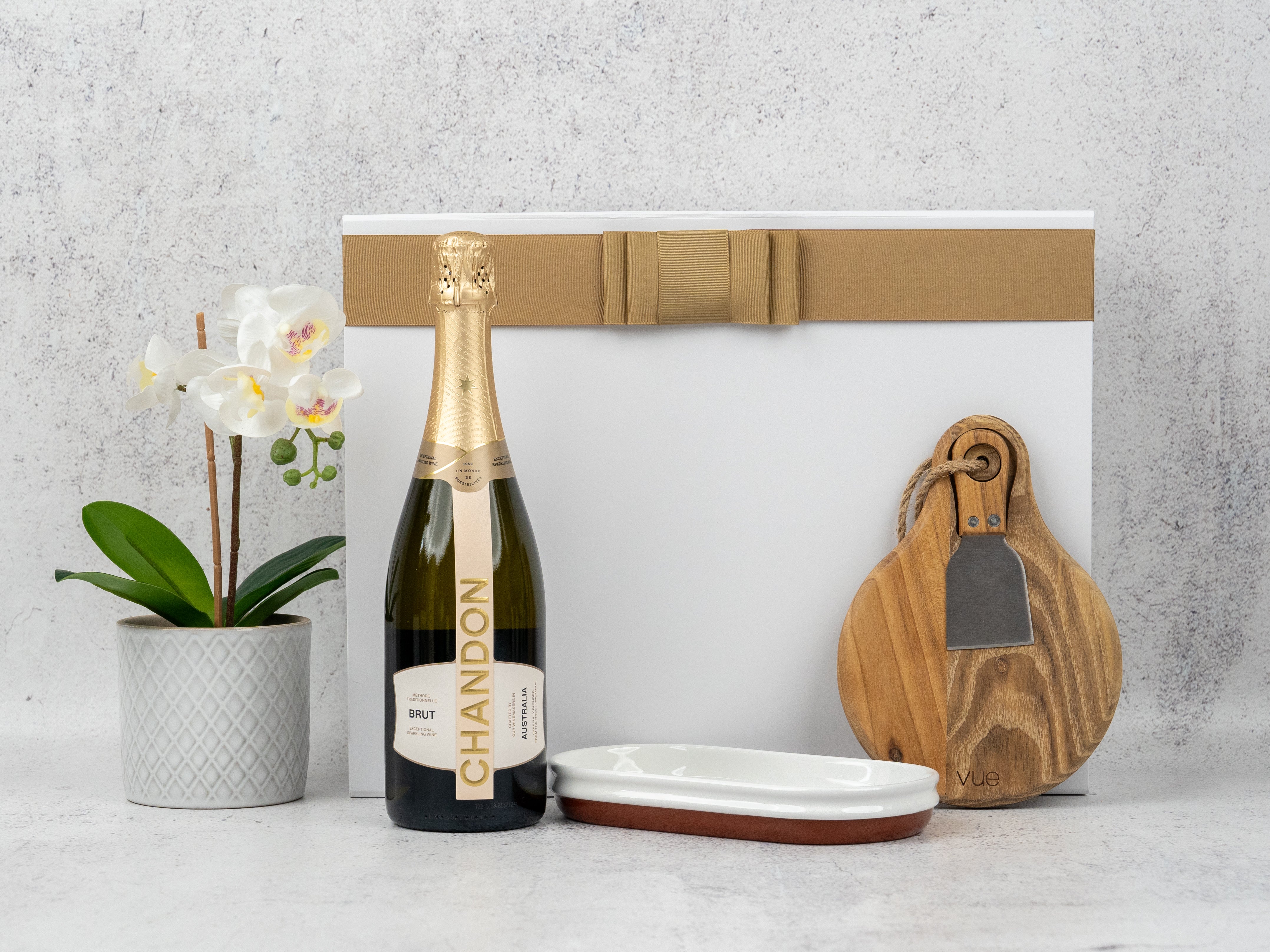 The Hamper Boutique Co’s Sparkling Celebration Hamper includes a bottle of Chandon Brut, white potted orchid, ceramic dish, two wooden cheese boards with a cheese knife in a gold-ribboned white box—all beautifully arranged.
