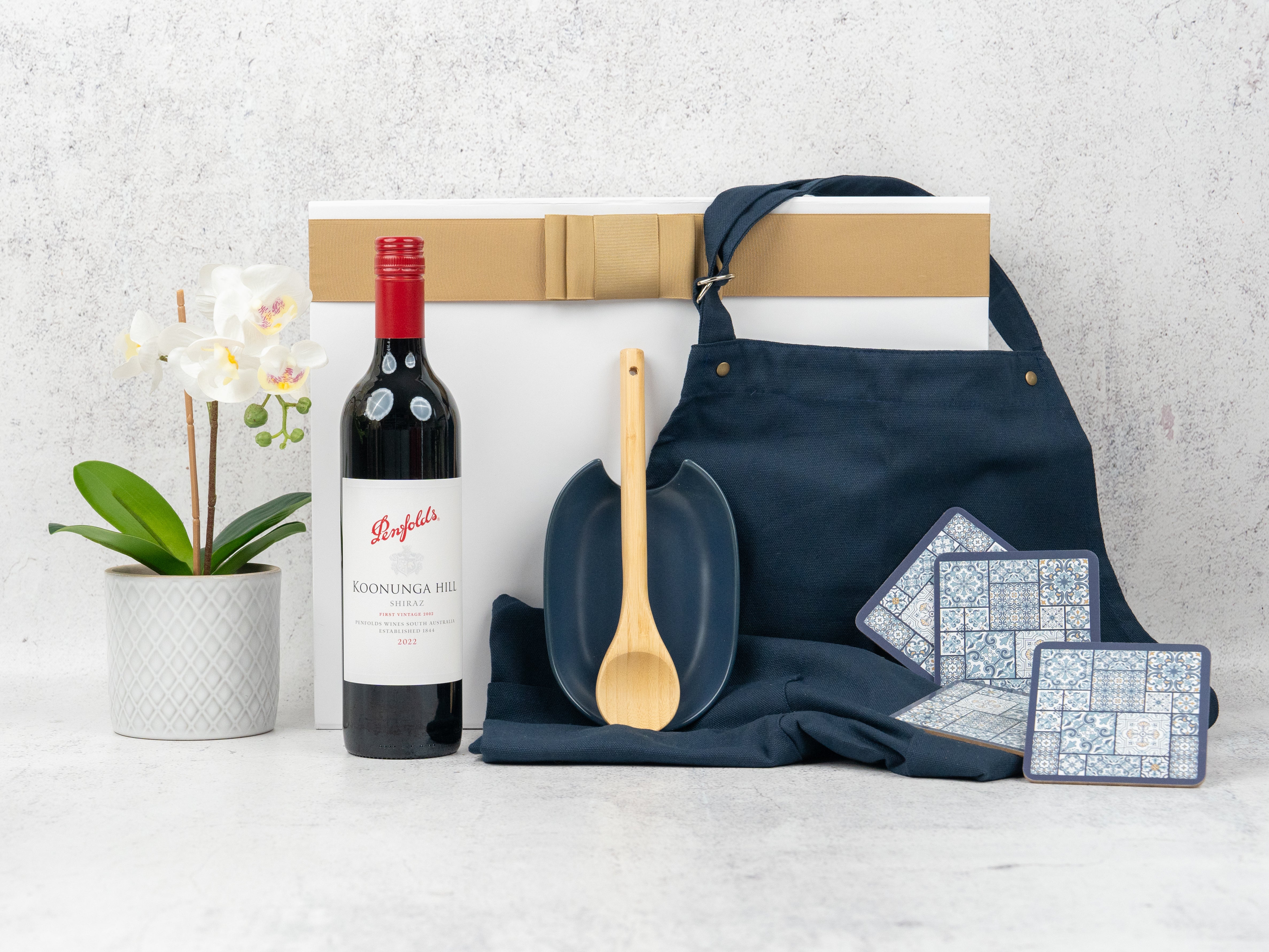 The Hamper Boutique Co’s "Wine and Dine" gift set features a potted white orchid, red wine, navy apron, wooden spoon, blue dish, patterned coasters, and a white gift box with gold ribbon.