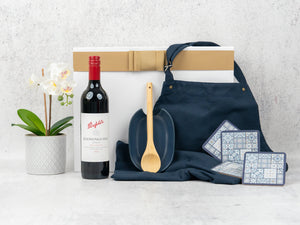The Hamper Boutique Co’s "Wine and Dine" gift set features a potted white orchid, red wine, navy apron, wooden spoon, blue dish, patterned coasters, and a white gift box with gold ribbon.