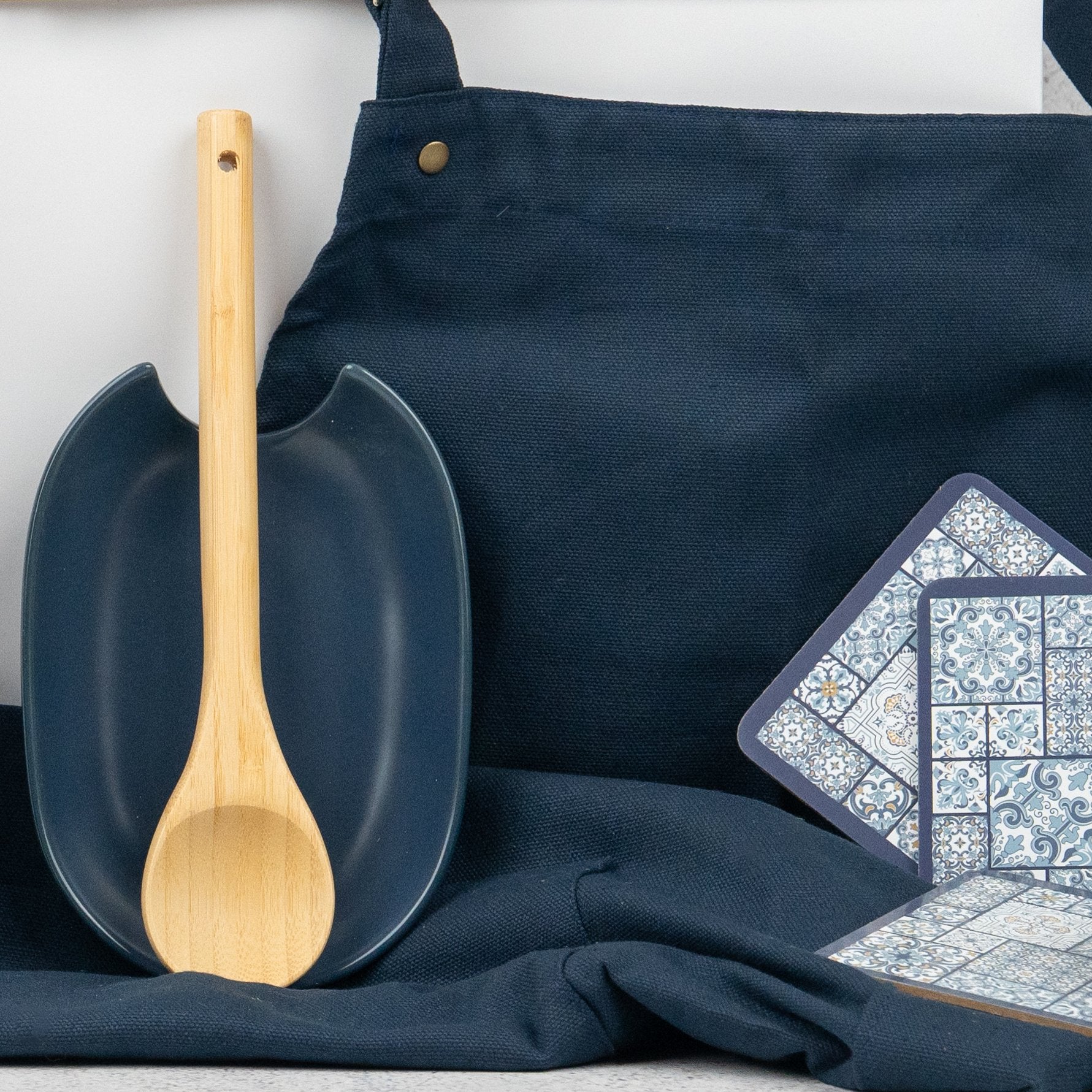 A wooden spoon rests on a dark blue spoon rest atop a matching apron from The Hamper Boutique Co's Wine and Dine set. Blue and white patterned coasters are arranged to the right, all against a neutral background.