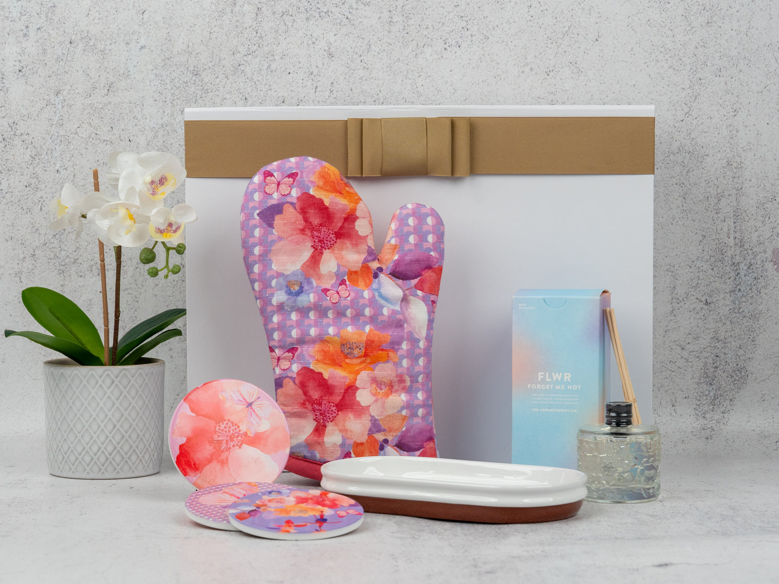 The Homeware Selection Hamper by The Hamper Boutique Co features a potted orchid, floral oven mitt, two coasters, rectangular dish, reed diffuser, and a white gift box with gold ribbon, all beautifully arranged on a light surface.