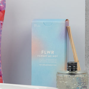 A pastel-colored Homeware Selection Hamper from The Hamper Boutique Co sits beside a glass reed diffuser with wooden sticks, with part of a multicolored fabric to the left. The background is light gray.