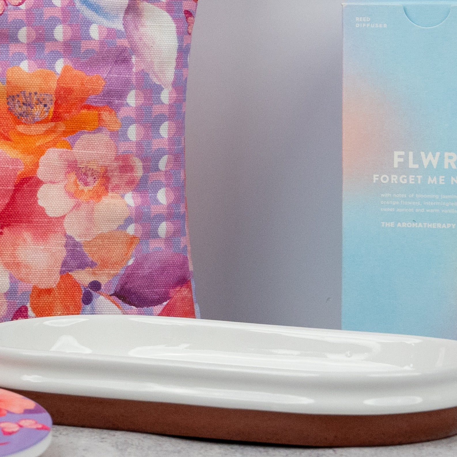 A close-up of a colorful floral-patterned pouch from The Hamper Boutique Co Homeware Selection Hamper sits next to a light blue FLWR FORGET ME NOT box and a white ceramic dish on a light surface.