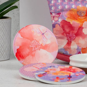 The Hamper Boutique Co’s Homeware Selection Hamper features three round coasters with vibrant floral designs in pink, orange, and purple, displayed on a white surface beside a patterned plant pot and a matching floral print bag.