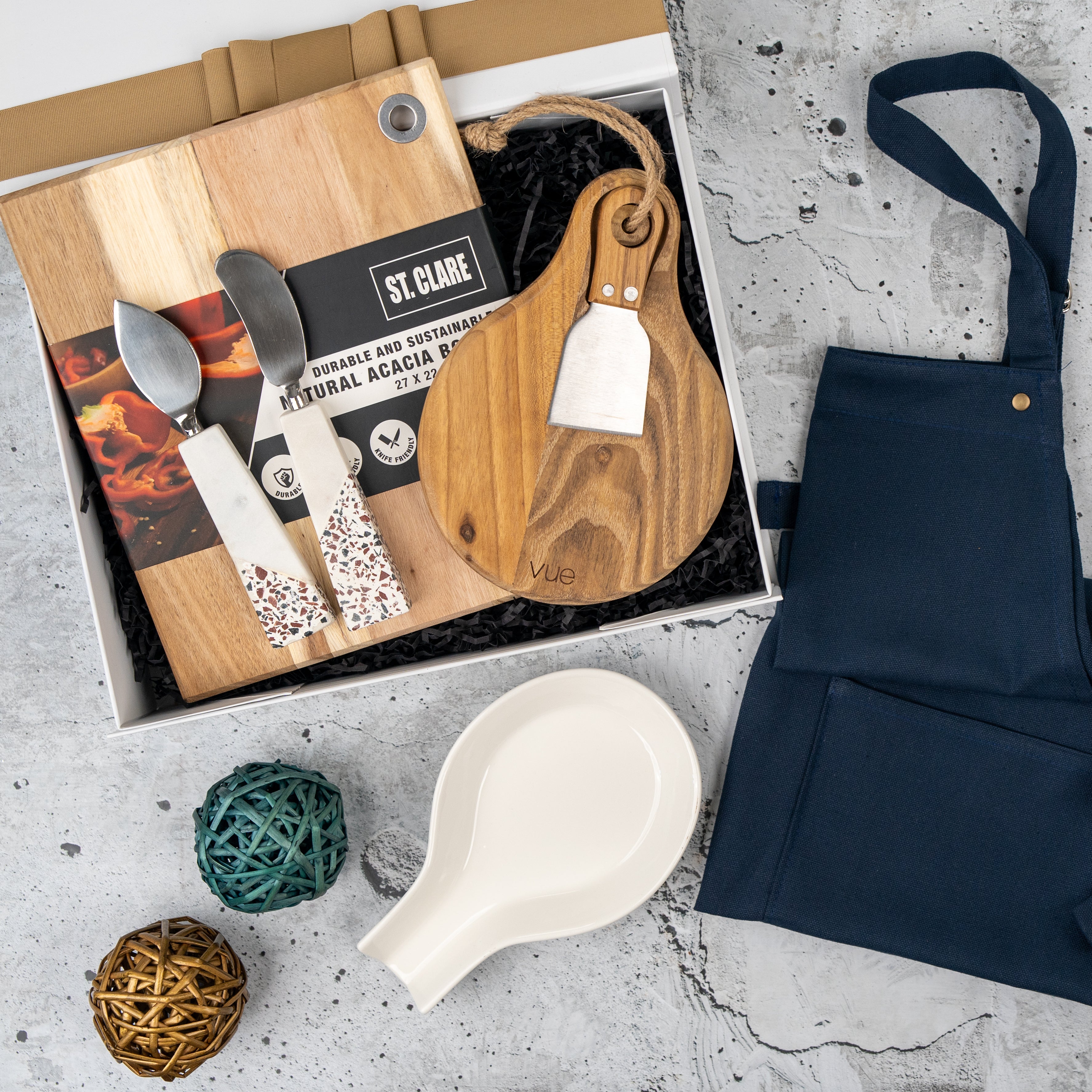 The Hamper Boutique Co’s Homeware Essentials Ensemble Hamper features a wooden cutting board, cheese board with spreader, marble-finish cheese knives, beige ceramic spoon rest, decorative balls, and navy apron on a gray textured surface.