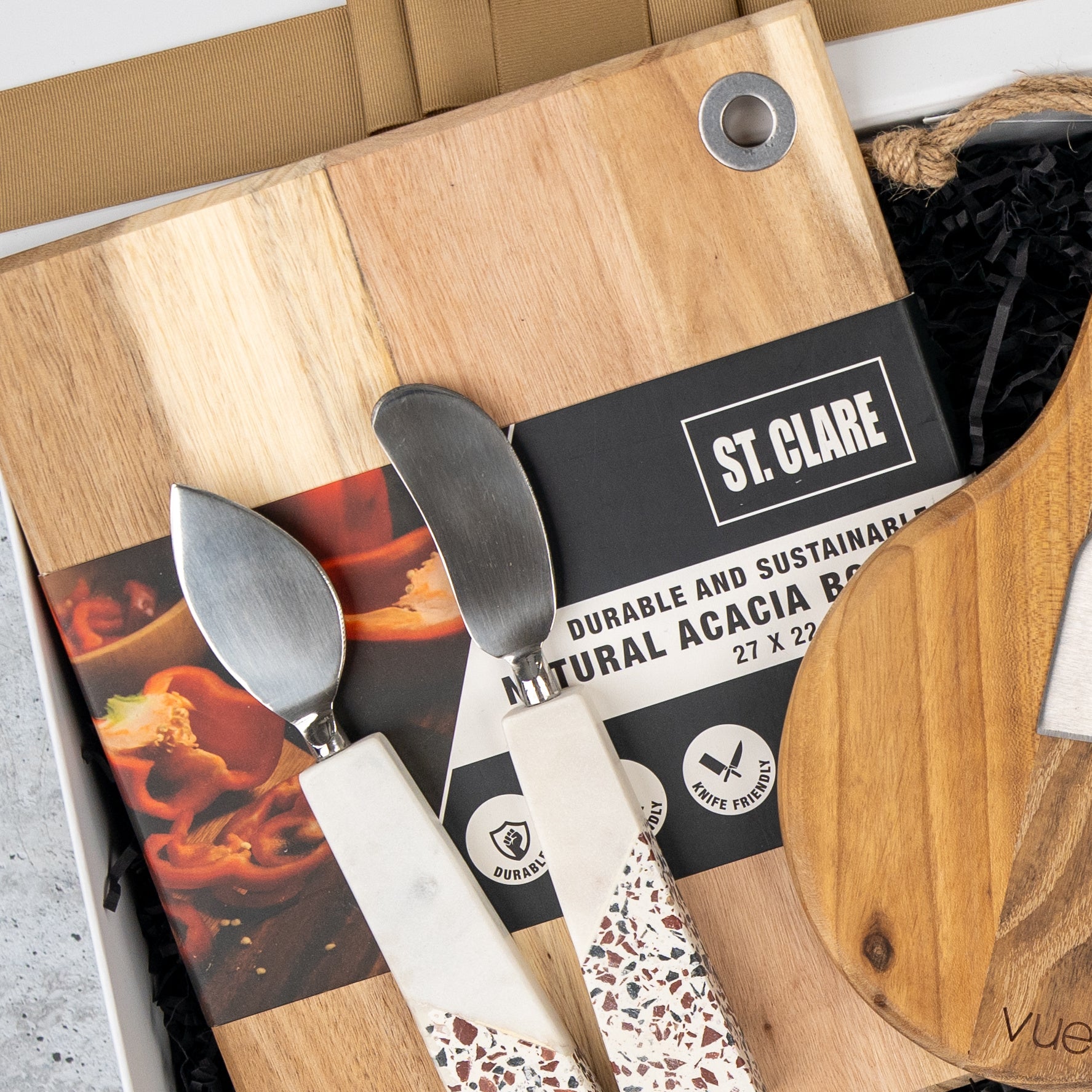 A close-up of The Hamper Boutique Co’s Homeware Essentials Ensemble Hamper, featuring a natural acacia wood cutting board and two cheese knives with decorative handles in branded packaging.