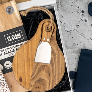 The Hamper Boutique Co’s Homeware Essentials Ensemble Hamper features a round wooden cheese board with rope loop, a short-handled cheese knife, and ‘vue’ engraving, presented on black shredded paper with partial packaging and a blue cloth nearby.