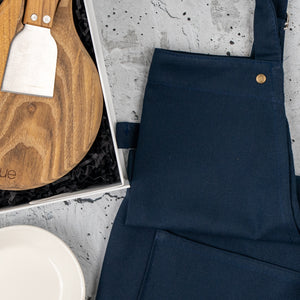 The Hamper Boutique Co’s Homeware Essentials Ensemble Hamper, featuring a navy apron, round wooden cutting board with cheese knife, and white plate, is beautifully displayed on a gray textured surface.