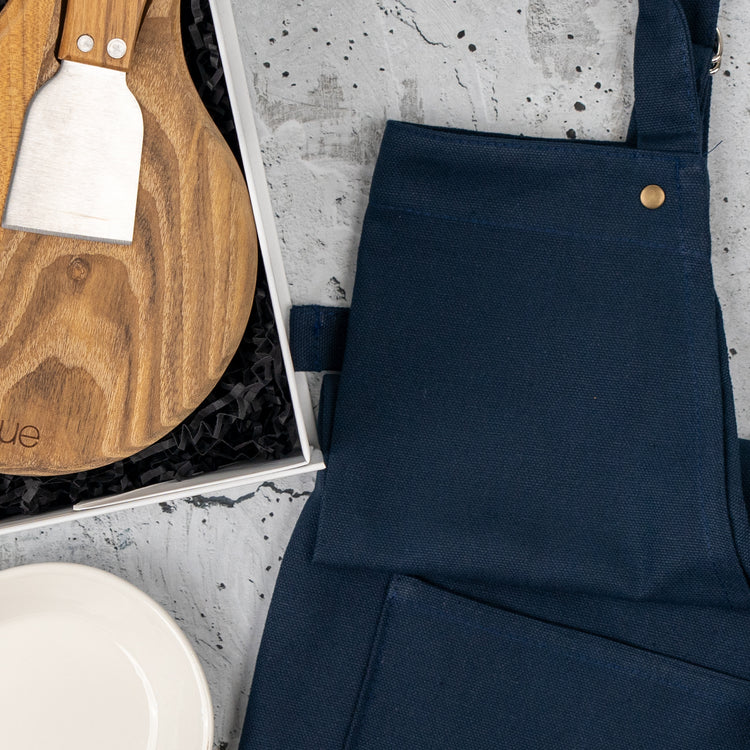 The Hamper Boutique Co’s Homeware Essentials Ensemble Hamper, featuring a navy apron, round wooden cutting board with cheese knife, and white plate, is beautifully displayed on a gray textured surface.