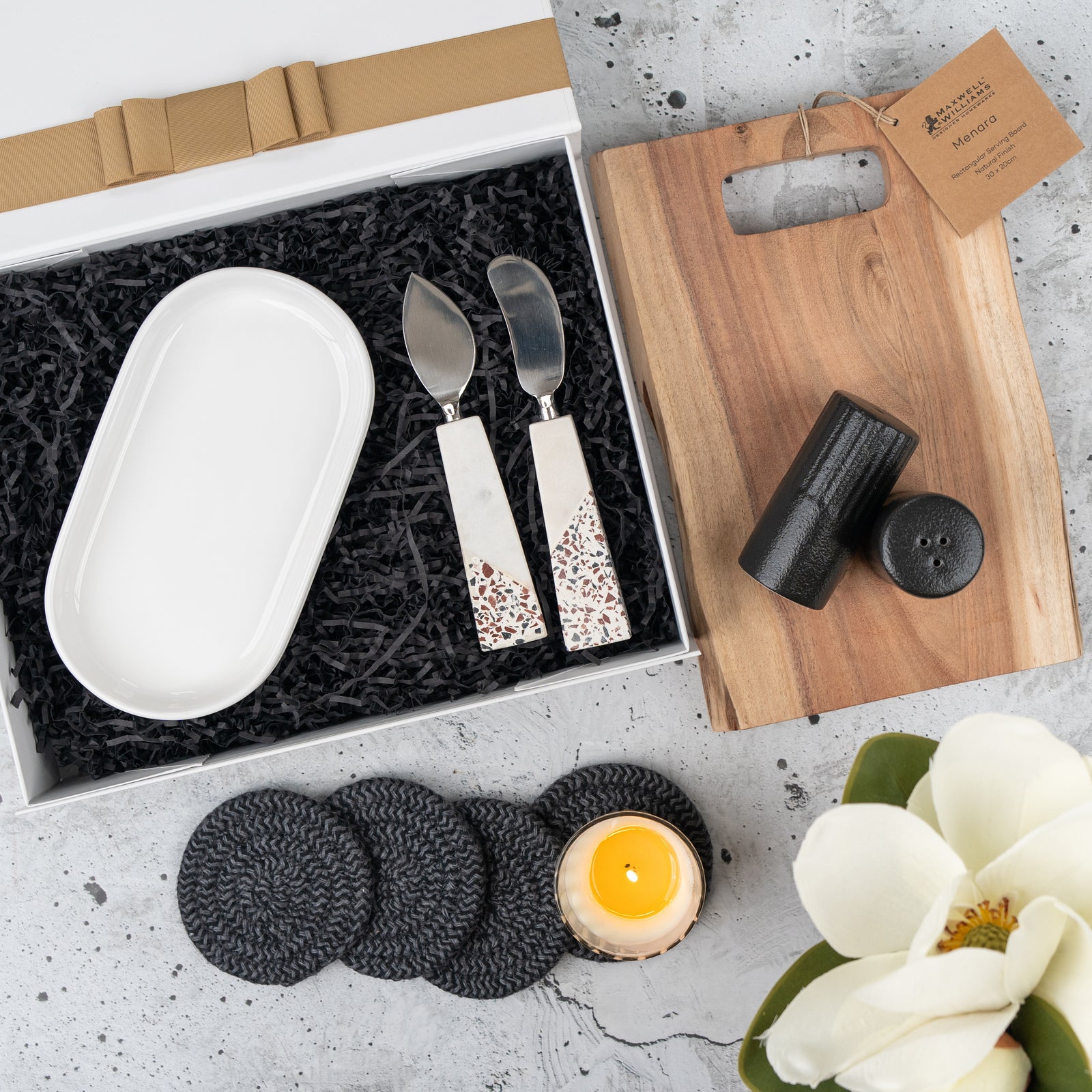 The Hamper Boutique Co’s Luxe Homeware Hamper features a white ceramic tray, two cheese knives, wooden board, black salt and shaker, four black coasters, a lit candle, and a white magnolia flower on a gray surface.
