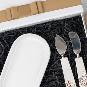 The Luxe Homeware Hamper by The Hamper Boutique Co features a white ceramic dish and two cheese knives with terrazzo-patterned handles, beautifully presented on black shredded paper and partially wrapped with a brown ribbon.