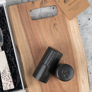 The Luxe Homeware Hamper by The Hamper Boutique Co features a rectangular wooden serving board with a handle slot, two black cylindrical salt and pepper shakers, and includes a terrazzo-handled knife, all displayed on a gray surface.