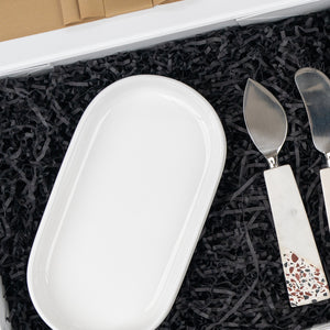 The Luxe Homeware Hamper by The Hamper Boutique Co features a white oval ceramic dish and two cheese knives with white and speckled handles, beautifully presented on black shredded paper inside the box.