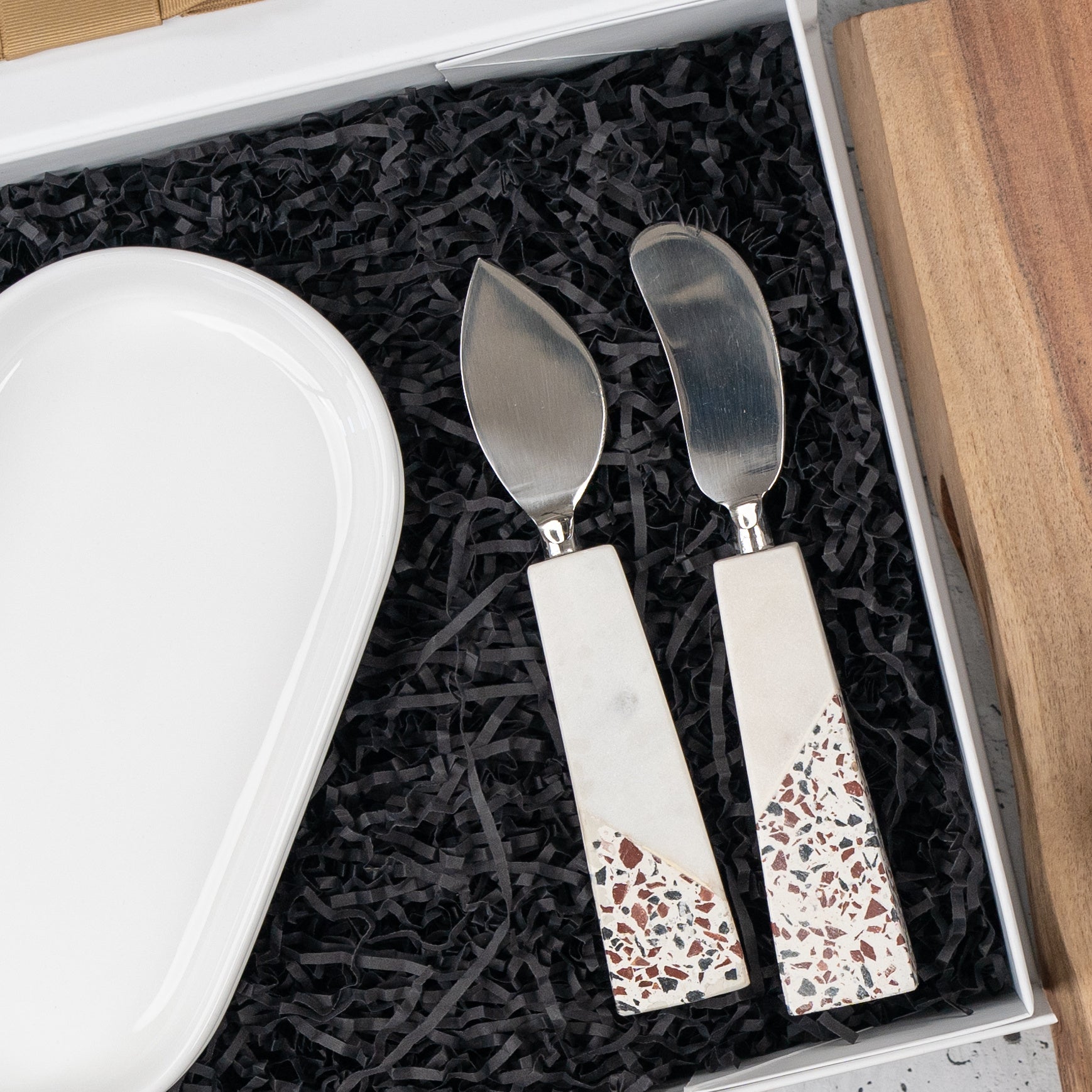 The Luxe Homeware Hamper by The Hamper Boutique Co features two cheese knives with terrazzo-patterned handles beside a white oval dish, all nestled in black crinkle paper inside a gift box.