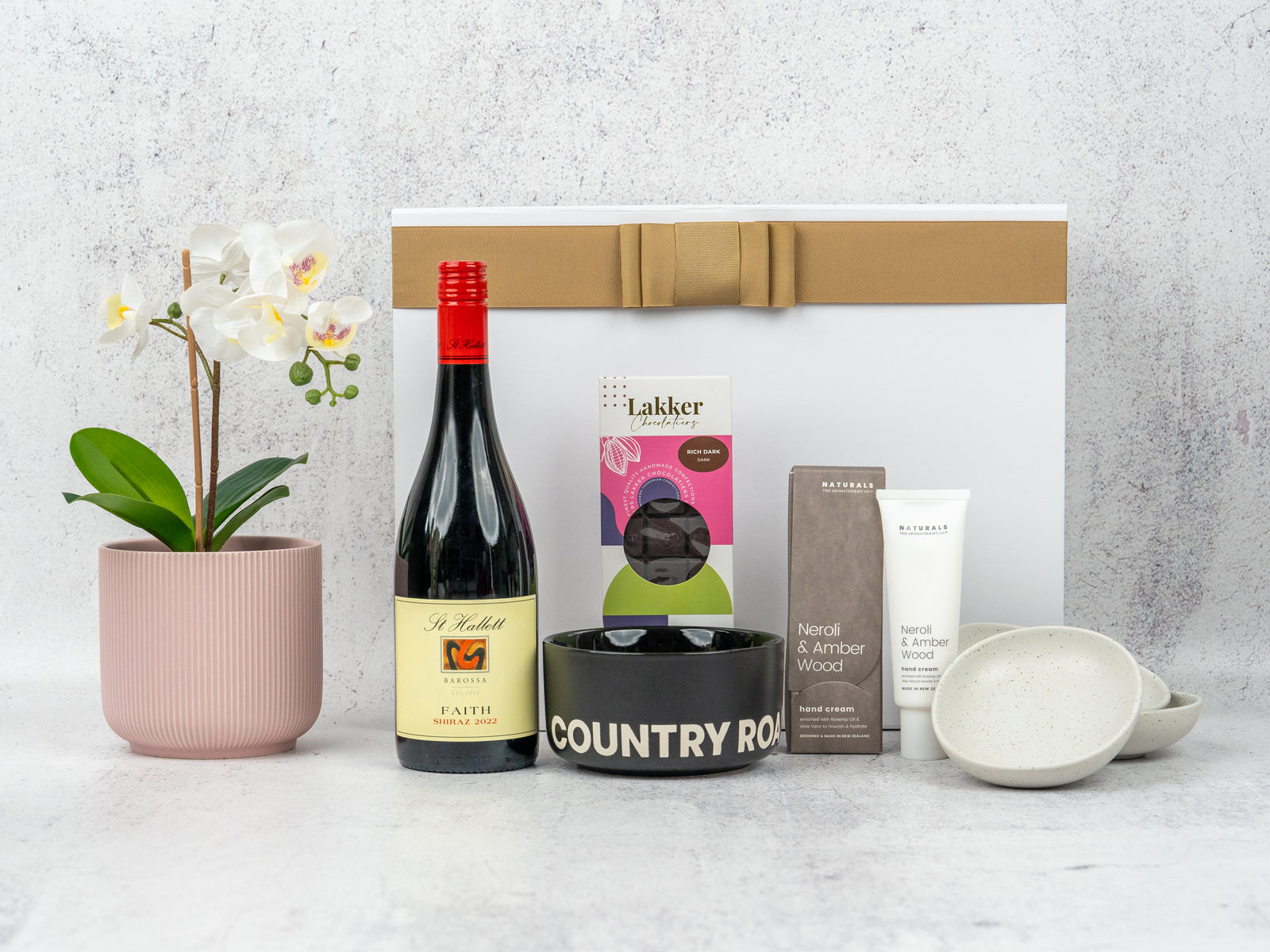 The Hamper Boutique Co's Wellness and Home Entertaining gift set includes a potted white orchid, red wine, chocolates, scented candle, hand cream, two small bowls, and a COUNTRY ROAD dish in a white box with brown ribbon.