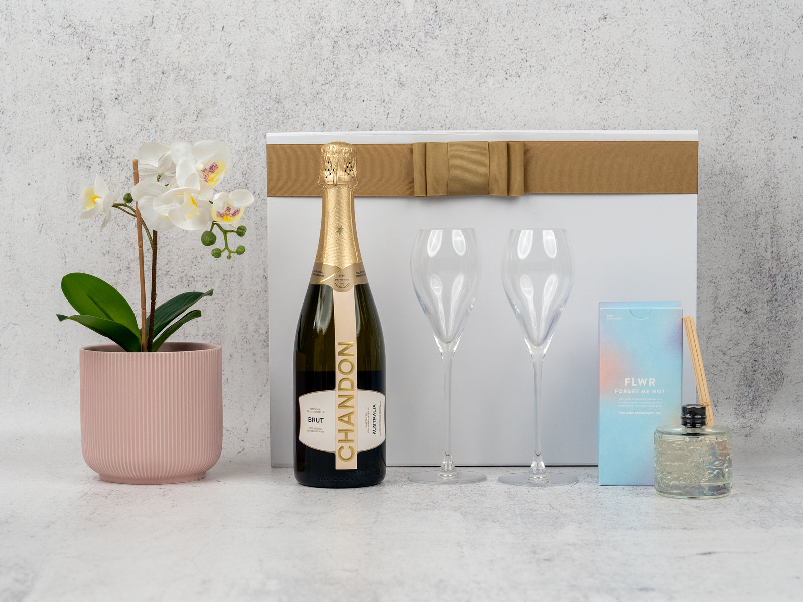 The Hamper Boutique Co’s Champagne Delight Hamper features Chandon sparkling wine, two wine glasses, a white orchid in a pink pot, a blue box, and a glass reed diffuser, all presented in a white box with a gold ribbon.