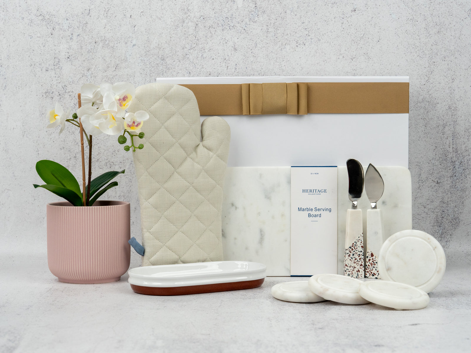 The Hamper Boutique Co's Prestige Home Entertaining Hamper features a potted white orchid, quilted oven mitt, marble serving board and coasters, serving utensils, and tray—all beautifully arranged on a light textured background.