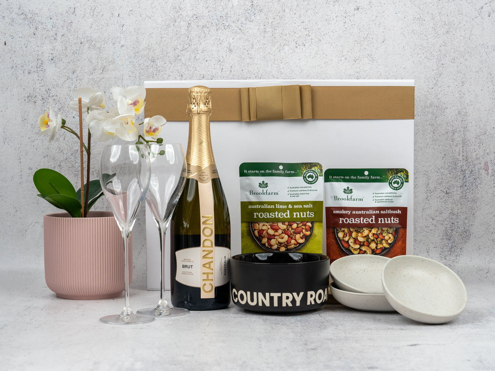 The Hamper Boutique Co’s Gourmet Sparkling Hamper includes a gold-and-white box, potted orchid, Chandon Brut, two champagne flutes, roasted nuts, black Country Road bowl, and two small white bowls—a sophisticated gift set.
