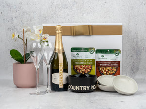 The Hamper Boutique Co’s Gourmet Sparkling Hamper includes a gold-and-white box, potted orchid, Chandon Brut, two champagne flutes, roasted nuts, black Country Road bowl, and two small white bowls—a sophisticated gift set.