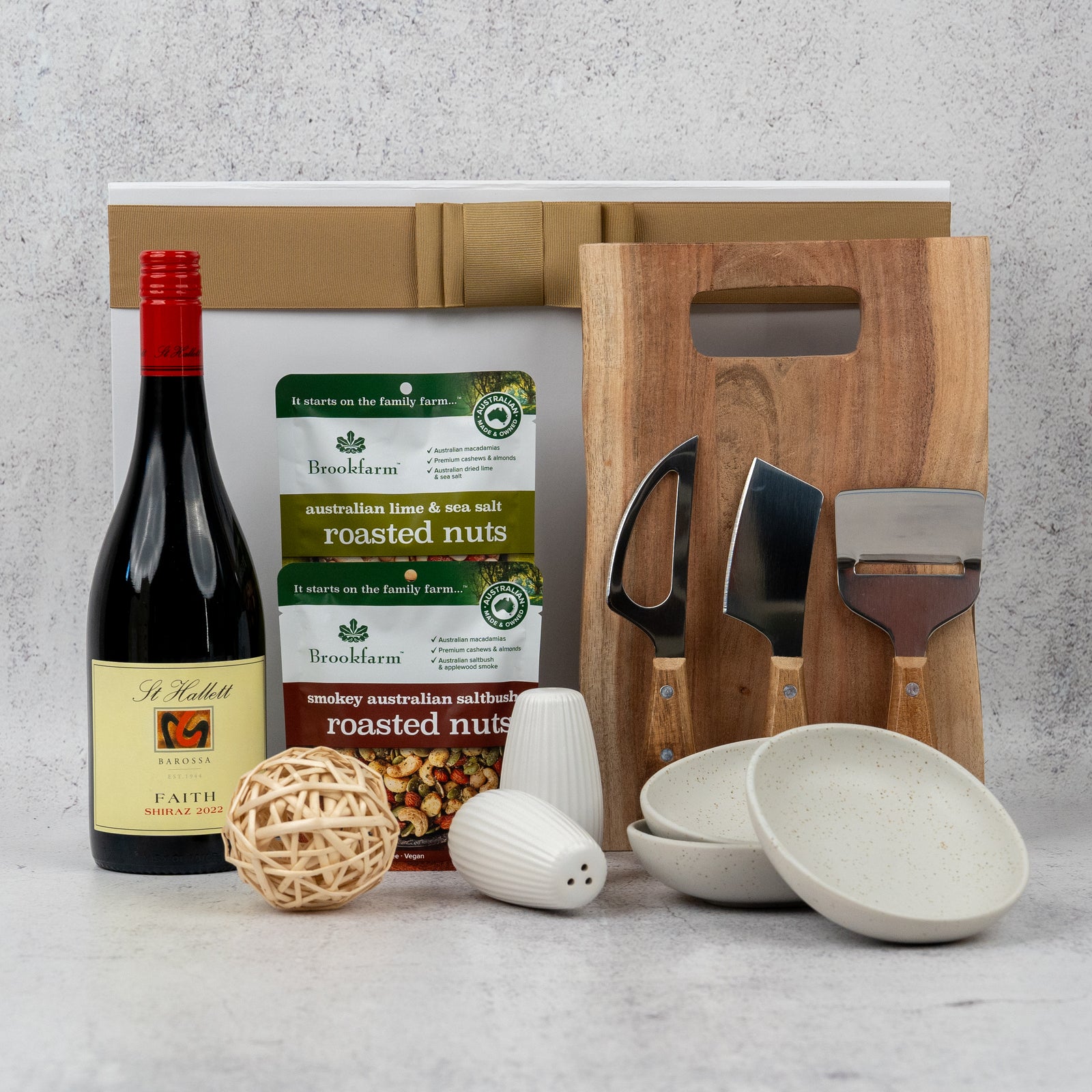 The Luxe Homeware and Shiraz Ensemble by The Hamper Boutique Co features red wine, roasted nuts, a wooden board, cheese knives, ceramic bowls, salt and pepper shakers, and a rattan ball—perfectly arranged on a light surface.
