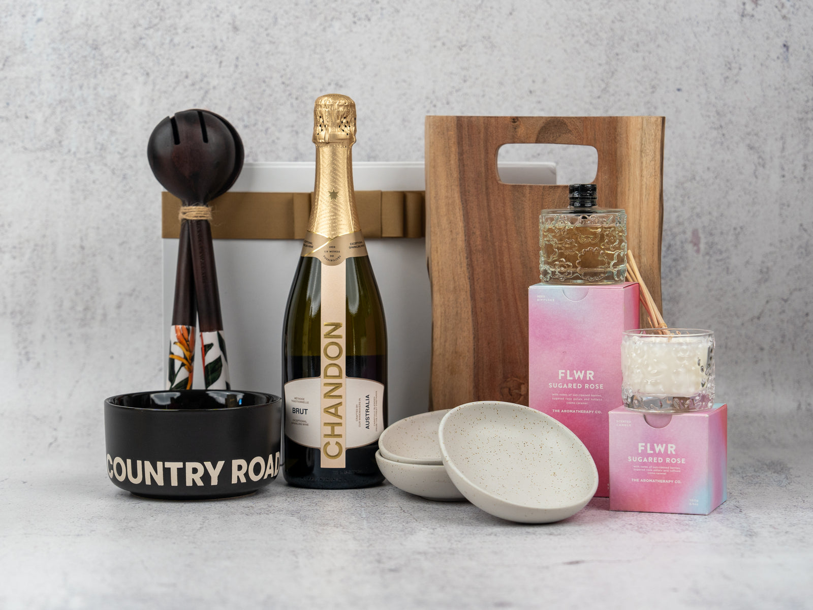 The Hamper Boutique Co’s Luxury Entertainer Hamper features Chandon champagne, two ceramic bowls, a wooden board, black Country Road mug, bath soap, candle, white box, and maracas—all beautifully arranged on a gray surface.