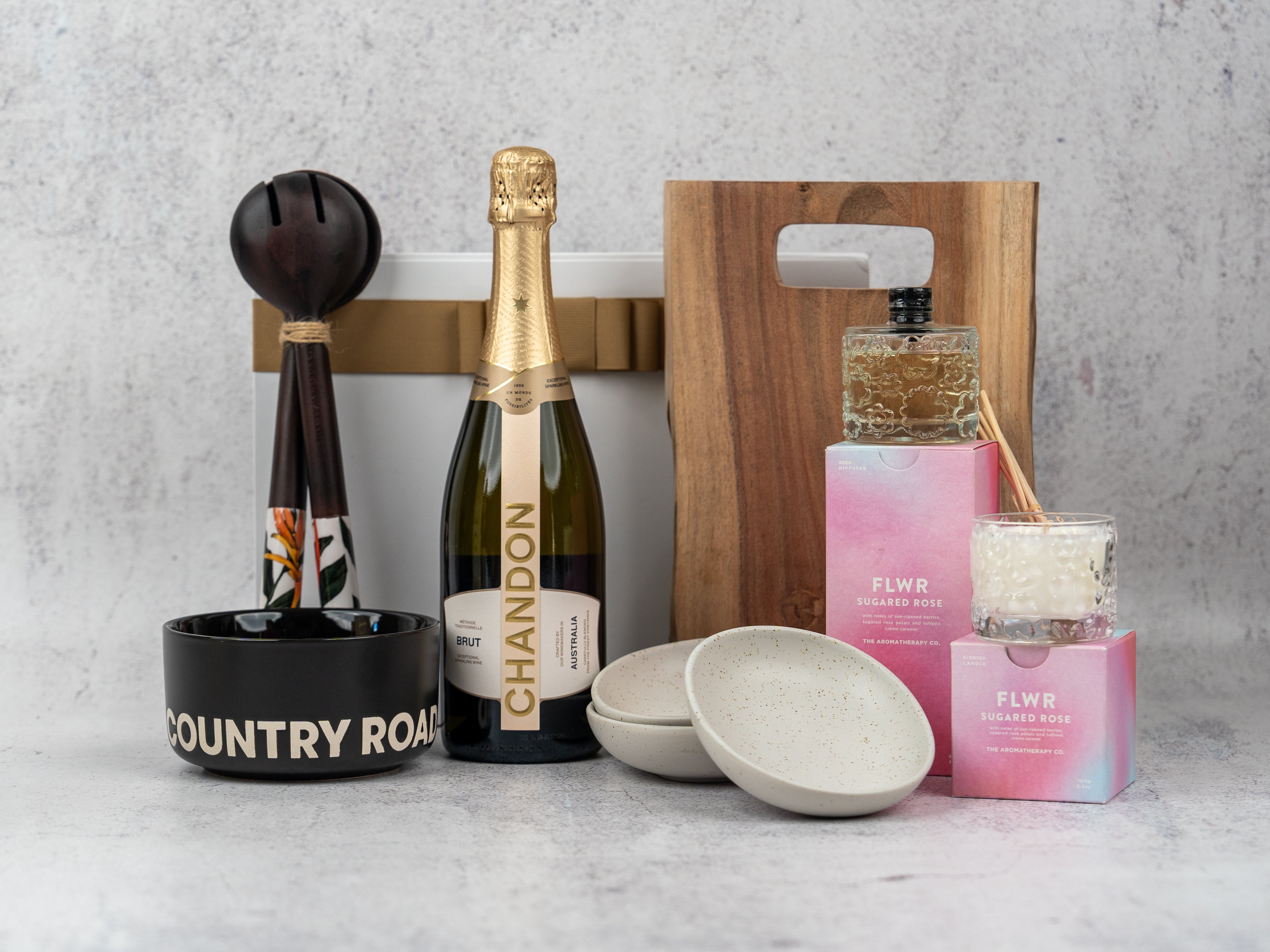 The Hamper Boutique Co’s Luxury Entertainer Hamper features Chandon champagne, two ceramic bowls, a wooden board, black Country Road mug, bath soap, candle, white box, and maracas—all beautifully arranged on a gray surface.