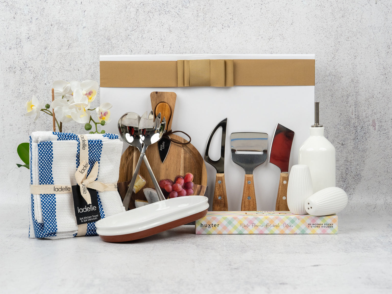The Hamper Boutique Co’s Homeware and Wellness Edition Hamper features kitchen essentials, such as a cheese board, knives, towels, ceramic olive oil dispenser, salt shaker, napkins, a small white vase, and a decorative artificial orchid.