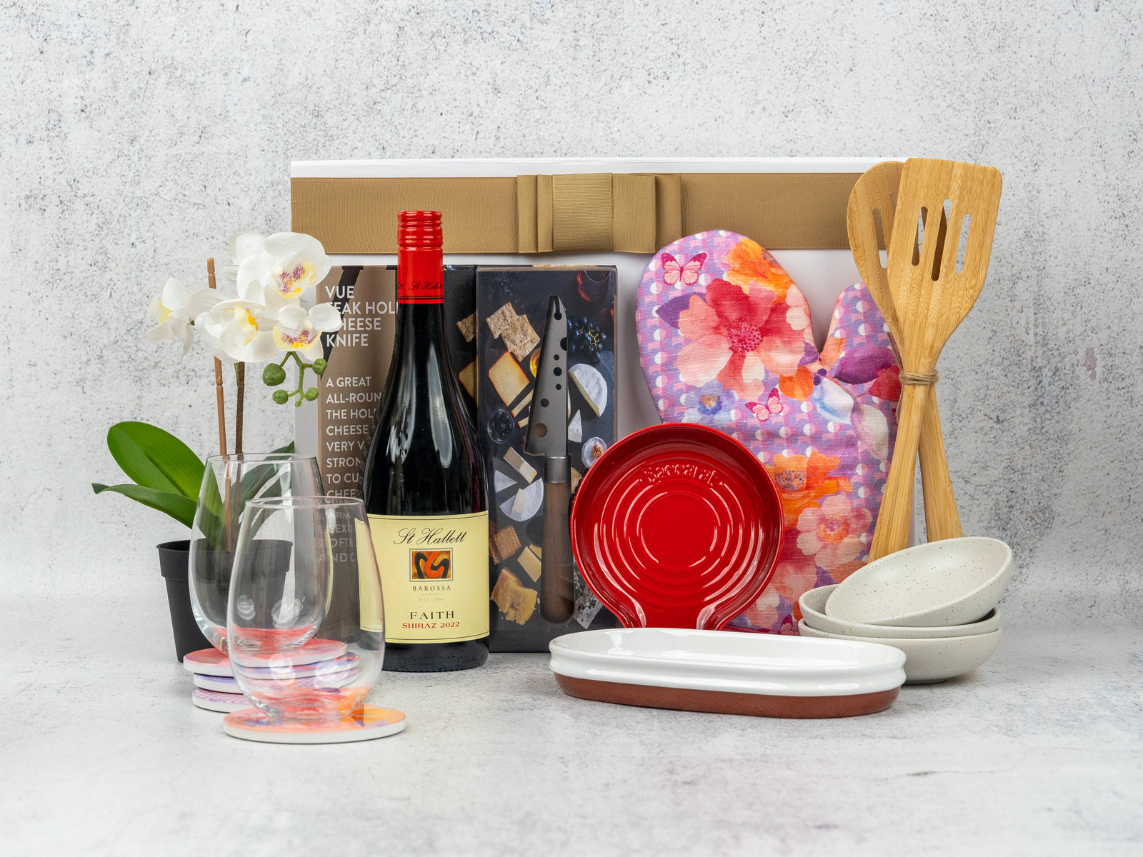 The Hamper Boutique Co's Wine Dine Gather set features red wine, two glasses, a white orchid, gourmet snacks, heart-shaped oven mitts, wooden utensils, ceramic dishes, and colorful coasters artfully arranged on a light surface.
