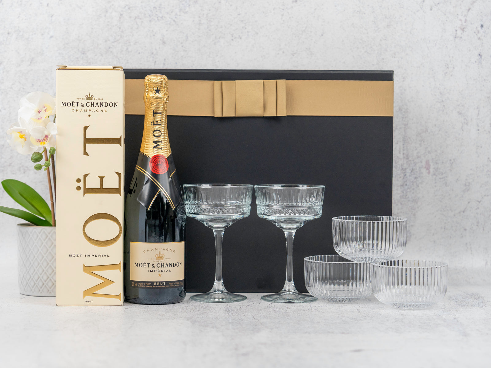 A Gin and Homeware Essentials Hamper by The Hamper Boutique Co is displayed on a light surface, featuring premium gin, two glasses, three serving bowls, a large black gift box with gold ribbon, and a potted white orchid.