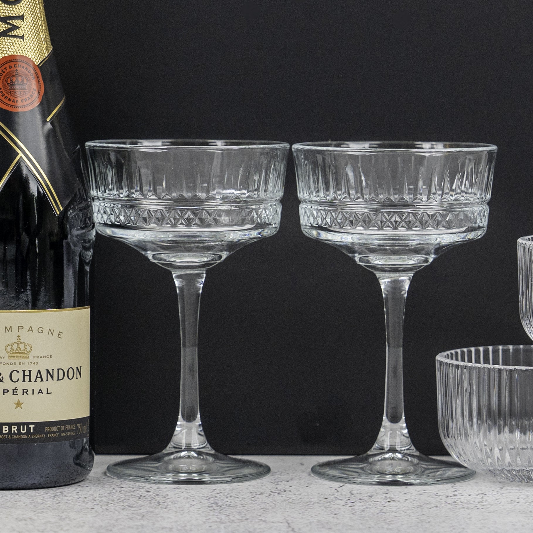 A Gin and Homeware Essentials Hamper by The Hamper Boutique Co sits beside two elegant, cut-glass coupe glasses on a light surface, set against a dark background.