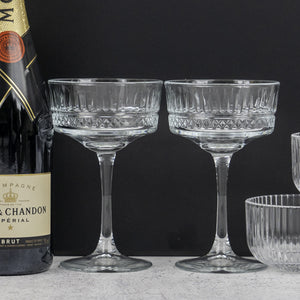 A Gin and Homeware Essentials Hamper by The Hamper Boutique Co sits beside two elegant, cut-glass coupe glasses on a light surface, set against a dark background.