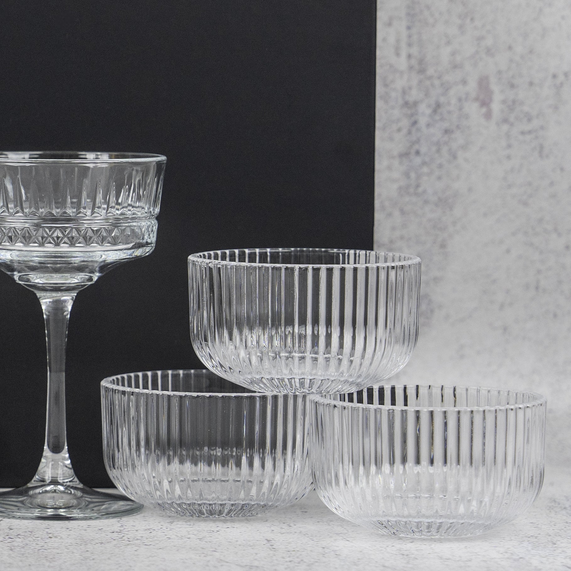 The Hamper Boutique Co’s Gin and Homeware Essentials Hamper features ribbed glass bowls and a matching stemmed glass with vertical ridges, elegantly arranged on a light surface against a black and gray background.