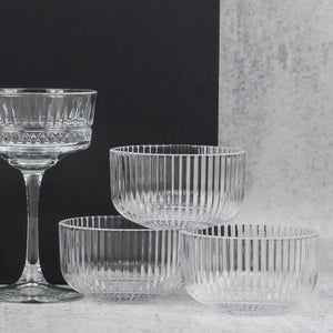The Hamper Boutique Co’s Gin and Homeware Essentials Hamper features ribbed glass bowls and a matching stemmed glass with vertical ridges, elegantly arranged on a light surface against a black and gray background.